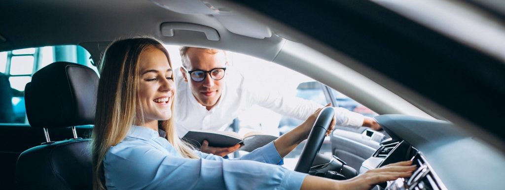 A smiling woman sitting in a car while a salesman explains the features.